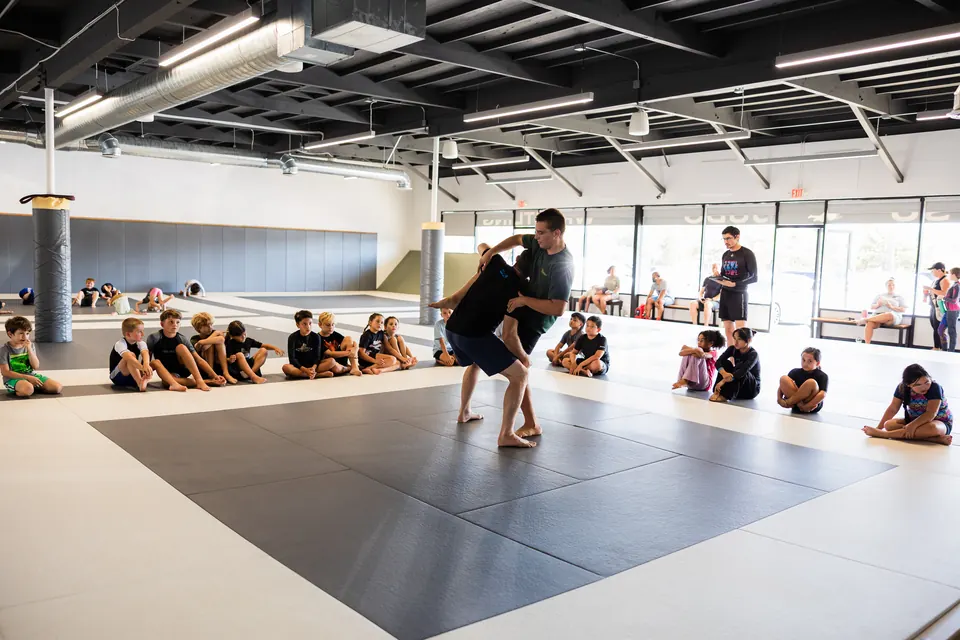 An instructor demonstrates a takedown while children sit around the mats
