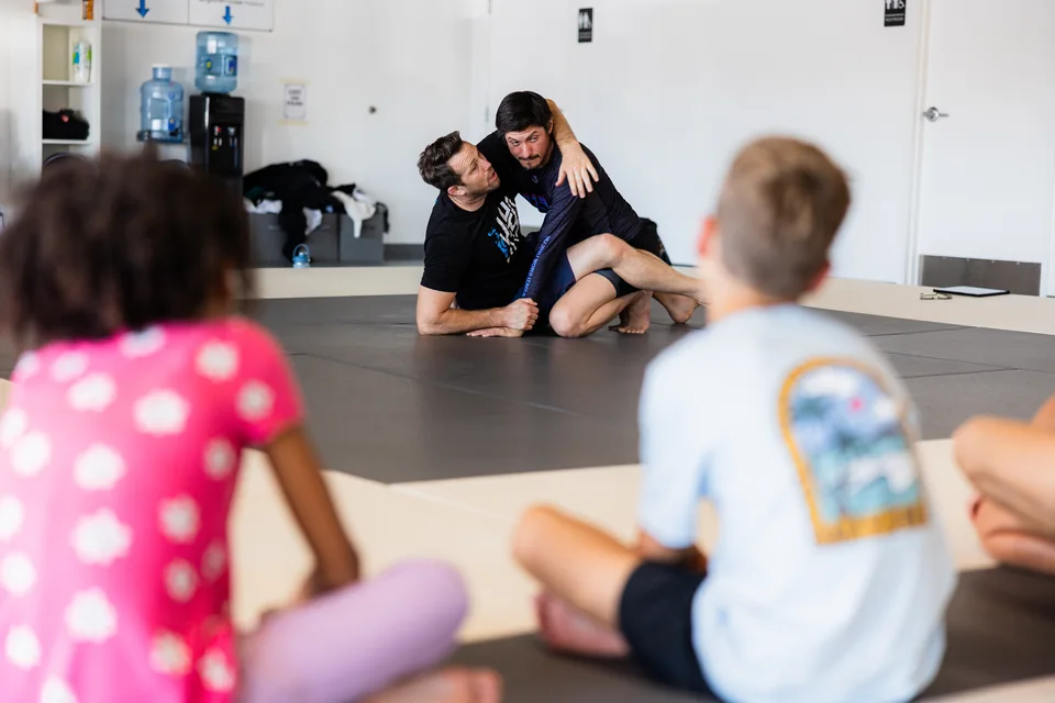 A coach demonstrates a grappling position while children watch