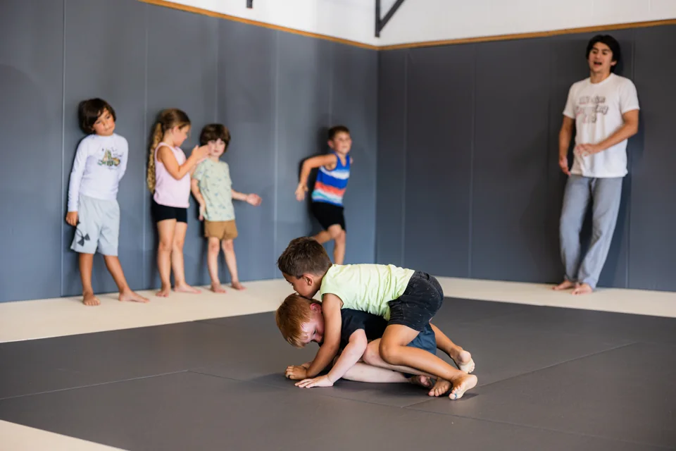 Two young children wrestle on the mats while Oscar Lopez watches