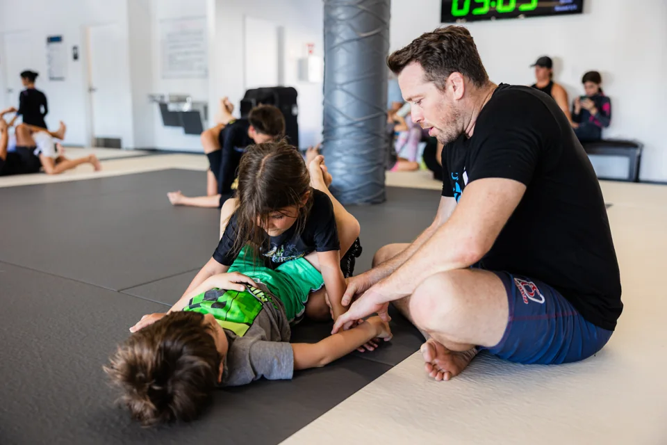 A coach kneels beside two children and guides their grappling practice
