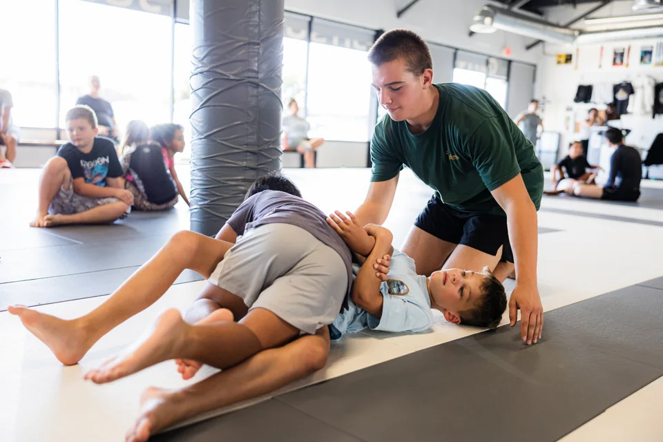 A coach kneels beside two boys grappling on the mats