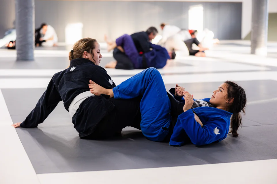 Two women grapple on the mats, one in a blue gi and one in a black gi