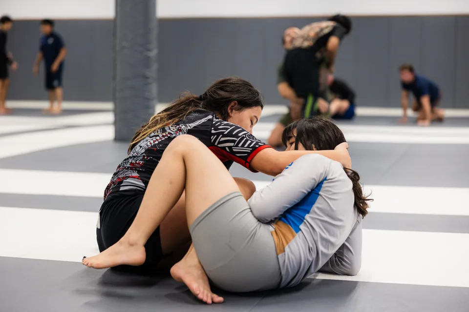 Two women practice a grappling position on the mats